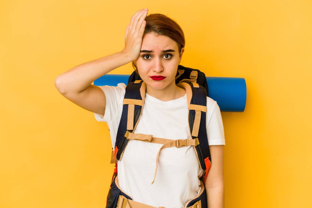 Young dark-haired woman looking disappointed with her hand on the side of her face and carrying a rucksack and rolled-up mat on her back.